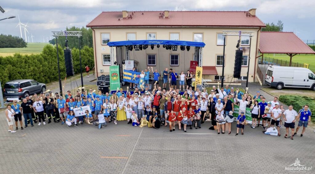 Grupa uczestników imprezy, w tym dzieci i dorośli, stoją razem na placu podczas grupowego zdjęcia podczas zakończenia ceremonii imprezy.