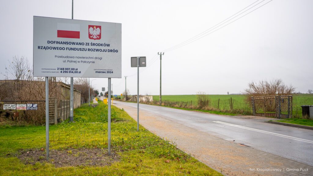 Tablica informacyjna dotycząca dofinansowania przebudowy ul. Polnej w Połczynie z widokiem na otwartą przestrzeń oraz nową nawierzchnię drogi.