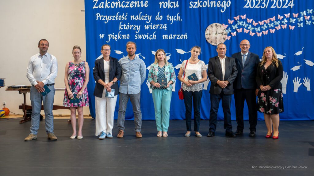 Grupa osób stojących na scenie przed niebieską dekoracją z napisem "Zakończenie roku szkolnego 2023/2024". Każda z osób trzyma nagrodę.