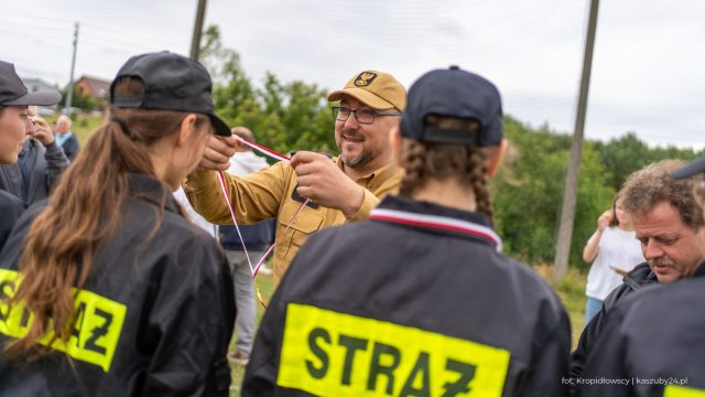Zdjęcie pokazuje mężczyznę w beżowym mundurze, który wręcza medal młodej strażaczce w czarnym mundurze. W tle znajdują się inni uczestnicy zawodów oraz obserwatorzy.