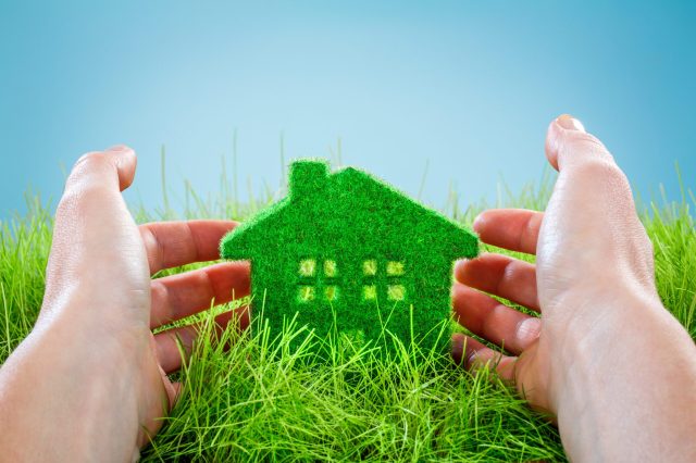 Eco House in green grass protected by the human hands on blue background.