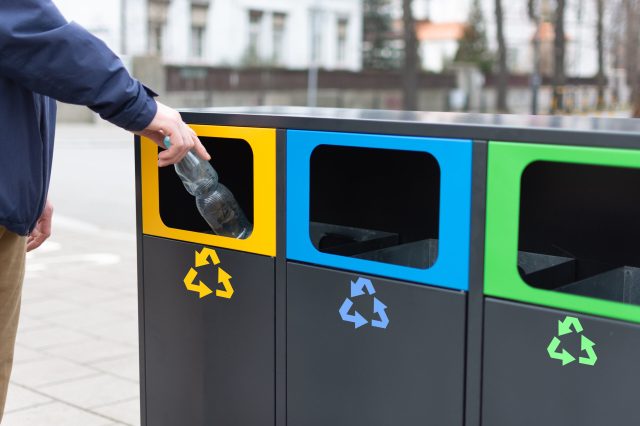 Hand of man throwing plactic bottle into trash bin for plastic waste. Modern colorful refuse bins for separate trash. Environmental consciousness lifstyle.