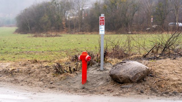 Czerwony hydrant przeciwpożarowy na tle pola i znak informacyjny dotyczący sieci wodociągowej.