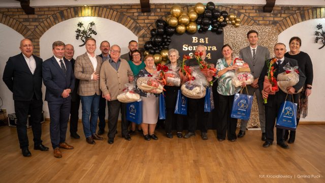 Grupa seniorów oraz zaproszeni goście pozują z nagrodami i kwiatami na tle dekoracji „Bal Seniora 2024”.