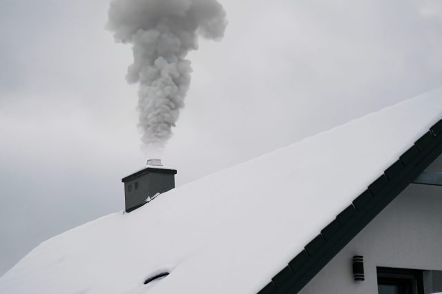 Smoke from a fireplace warms up a house in a winter wonderland as snow falls and covers the roof and the surrounding trees.